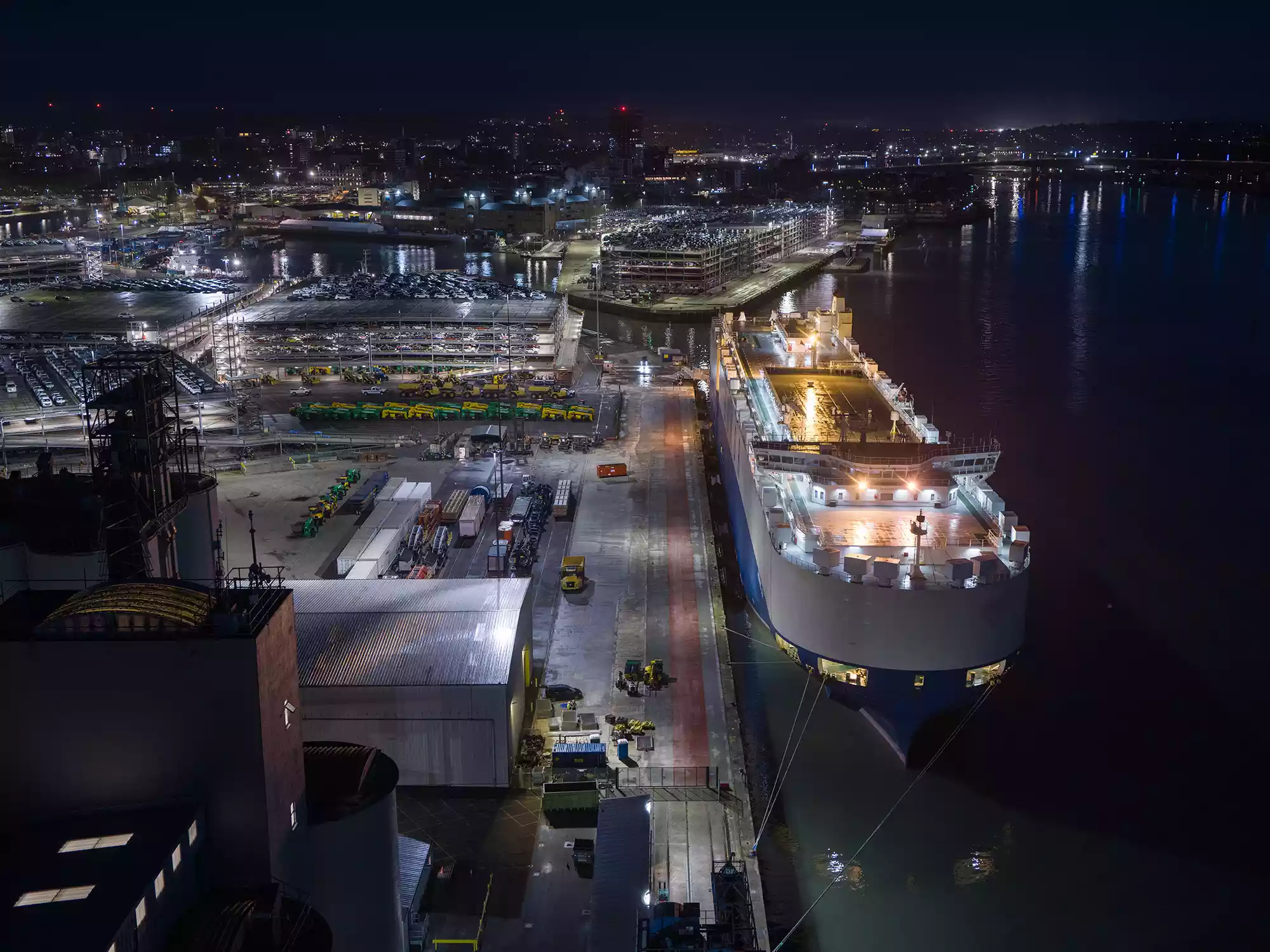 Aerial night view of a cargo ship moored beside an illuminated port terminal with vehicle storage decks